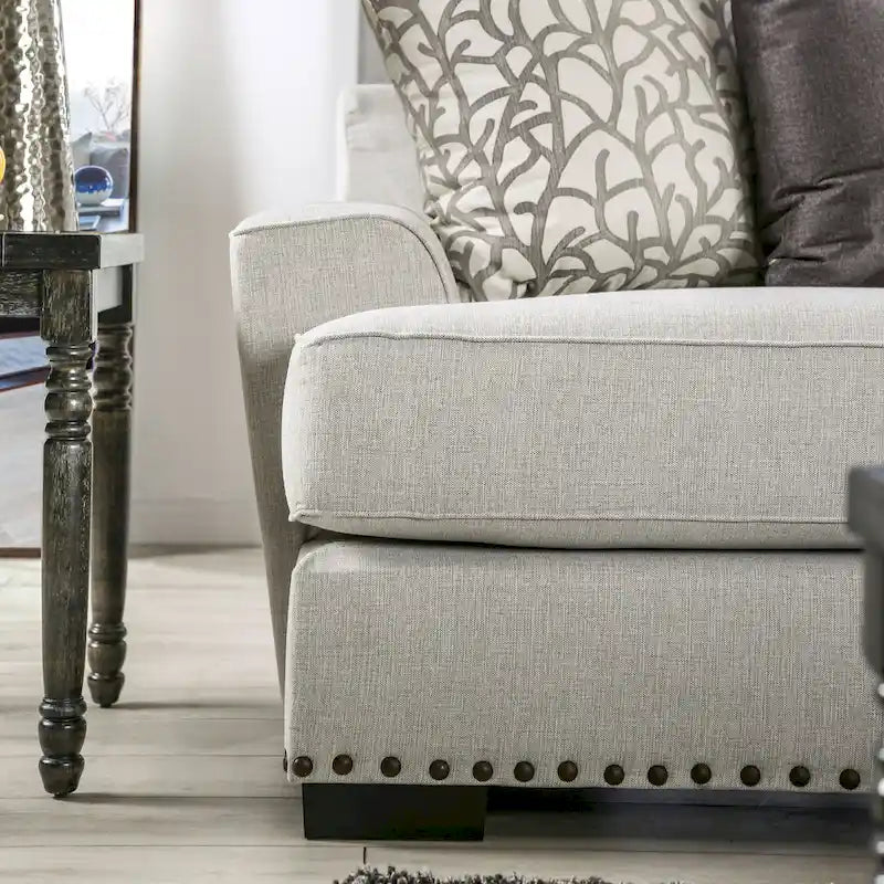 Sofa z jasnoszarą tapicerką materiałową w stylu przejściowym, ozdobiona listwą z gwoździami tapicerskimi i poduszkami dekoracyjnymi
